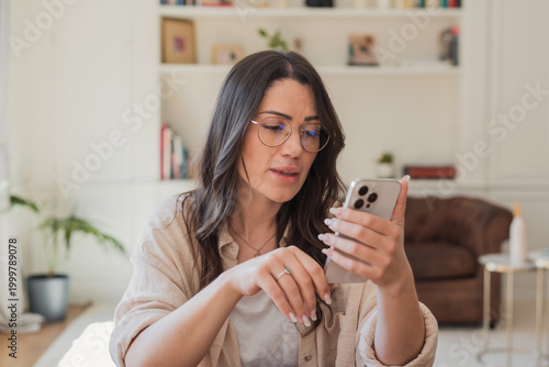 Obraz na plátne A woman in glasses uses her smartphone in a bright living room, focused on a video call or messaging