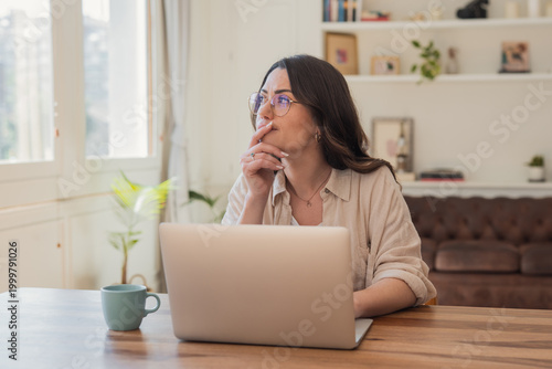 фотографія Young woman looks thoughtful while working on a laptop in a bright, cozy home office; relaxed remote work atmosphere with natural light, coffee mug, and comfortable living room background