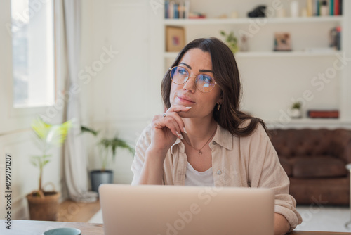 A thoughtful woman in glasses sits at her laptop in a bright, cozy home office, gazing thoughtfully while working remotely, studying online, planning projects and brainstorming ideas.