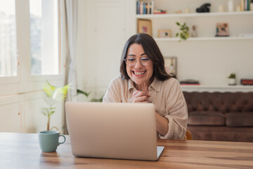 A joyful woman in glasses smiles and clasps her hands during a laptop video call, working from a cozy home office with plants, a mug and a comfortable sofa in the background.