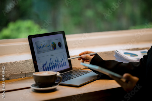 Professional woman working on laptop in café, enjoying hot drink, reviewing charts, planning projects, and staying focused in modern business lifestyle.
