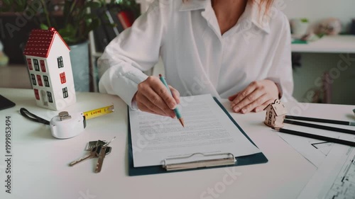 Female real estate agent meeting client in modern office, reviewing property contract and pointing where sign. Home purchase consultation with house model, floor plan and mortgage paperwork concept.