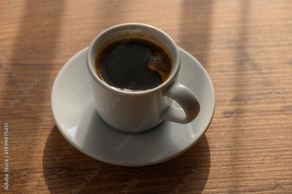Fototapeta premium A steaming cup of coffee on a wooden table with morning light streaming in