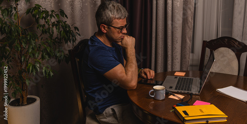 Tired freelancer drinking coffee while working late at night on laptop at home