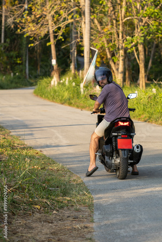 A man wearing a helmet and casual clothes sits on a black scooter on a sunny rural road in Asia, looking back over his shoulder, with lush greenery and trees along the scenic route.