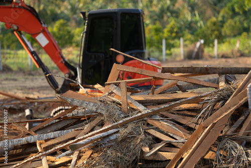 An orange excavator stands behind a large pile of wooden debris and palm tree remains on a sunny day. This image captures the process of land clearing and waste management in a natural setting.