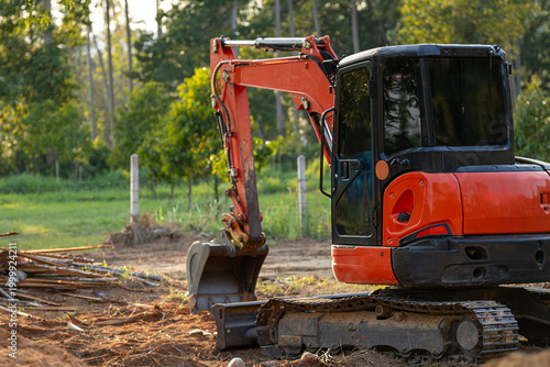 A vibrant orange and black mini excavator stands ready on a dirt construction site, with a pile of wooden debris nearby. 