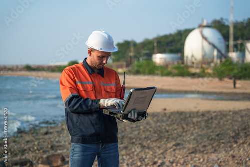 Male engineer in hard hat using rugged laptop for remote site inspection near gas storage tanks. Professional technician monitoring industrial energy infrastructure at coastal plant facility