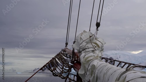 A static shot from the bow of a traditional sailing vessel as it navigates the icy waters of Antarctica's Danco Coast, featuring a wooden bowsprit and snow-covered mountains in the distance