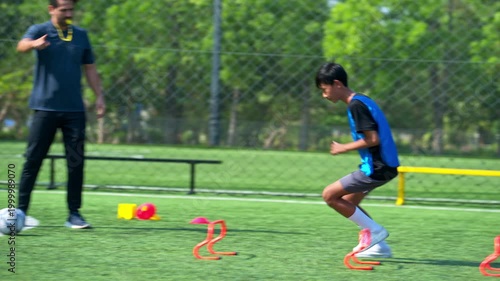 International school student running agility drill over hurdles during soccer practice, supervised by coach on field
