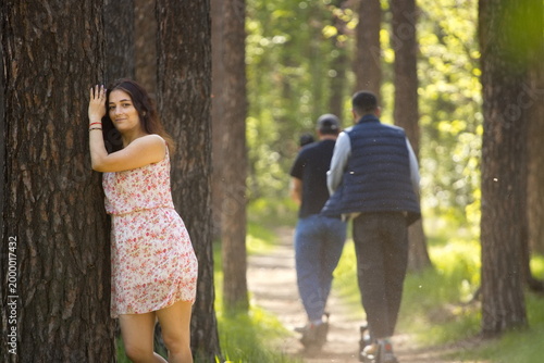 A young woman walking in the forest. A beautiful woman resting in a forest, leaning against a pine tree trunk. Behind her, a group of guys on scooters rides along a path.