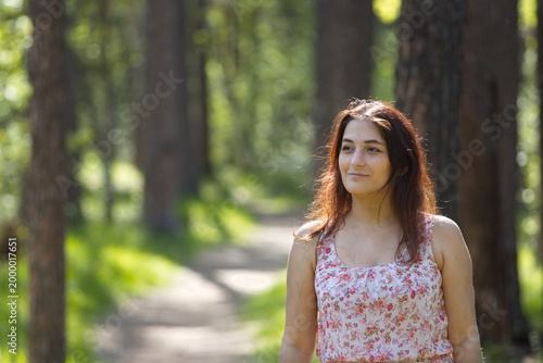Portrait of a young woman in a park. A woman stands on a path between pine trees and looks away. The background is beautifully blurred.