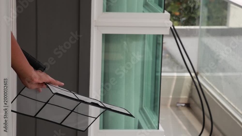 Hand holding a clean air conditioner filter and using an electric blower to remove water droplets after washing near a balcony window.