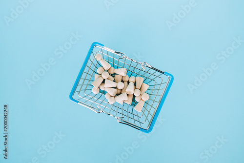 Small wooden peg dolls fill miniature wire shopping basket on bright blue surface. Cost of living crisis, inflation impact on families, basic needs affordability, Consumer purchasing power.
