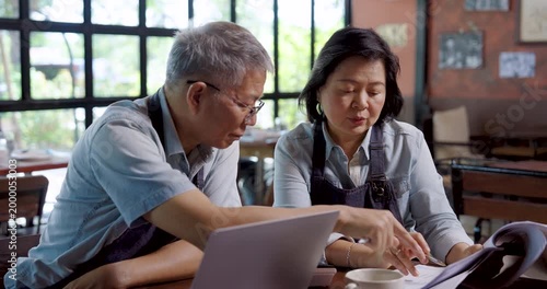 Asian senior couple cafe owner wearing aprons sitting at wooden table analyzing financial documents with laptop, teamwork management showing cooperation discussion problem solving in family restaurant