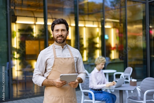 Foto A smiling man wearing an apron holds a tablet while standing outside a restauran