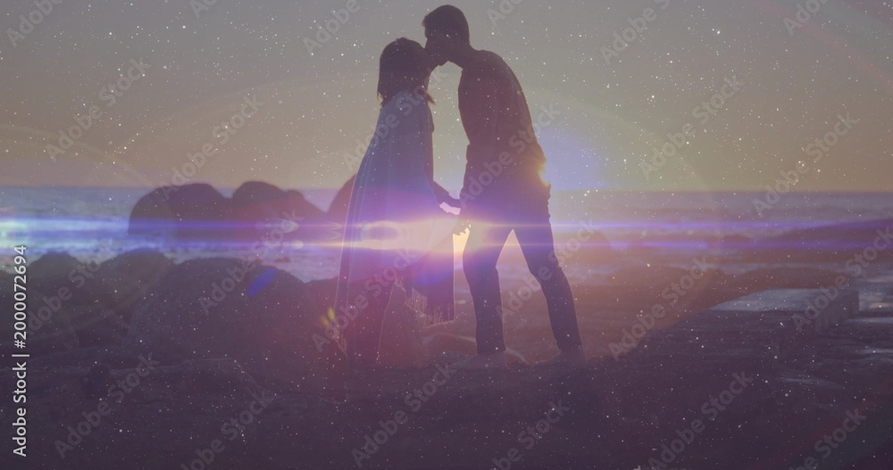 Naklejka premium Holding hands, couple in flowing dress and trousers touching on rocky beach at sunset, lens flare