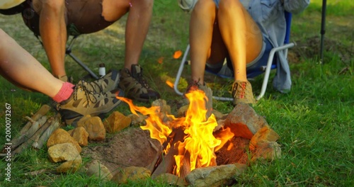 Sitting three adults warming feet by campfire on grassy lawn, with folding chairs and hiking shoes