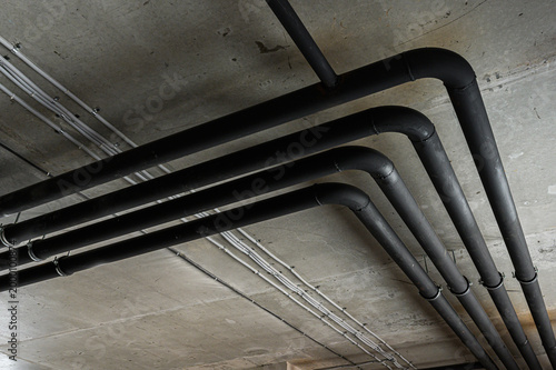Schilderij op canvas Black insulated pipes run along a concrete ceiling, secured with metal clamps; alongside are white electrical conduits, creating an industrial look