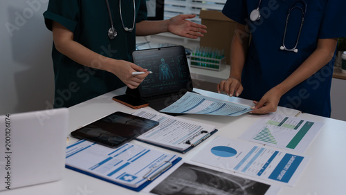 Medical team in scrubs reviewing patient charts and lab reports on desk with laptop showing digital anatomy scan in hospital office.