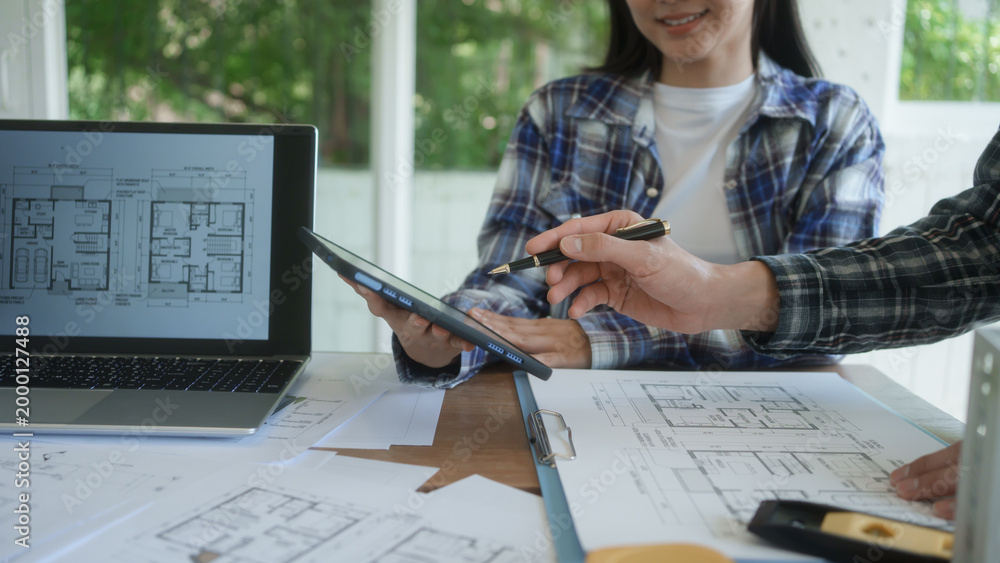 Fototapeta Architects reviewing house floor plans at desk, laptop and tablet in bright office, collaborating on home design project with blueprints and documents.