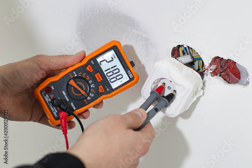 Photos Close-up of a professional technician's hands using a digital multimeter to check the voltage in a power socket during home electrical repair or maintenance