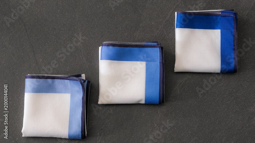 Trio of elegantly folded white handkerchiefs featuring various shades of blue border patterns and contrasting hand-rolled edges on a dark textured background.
