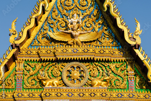 Close-up of a traditional Thai temple roof with intricate golden carvings, featuring a Garuda figure, Dharma wheel, and deer, set against a clear blue sky, highlighting ornate architecture.