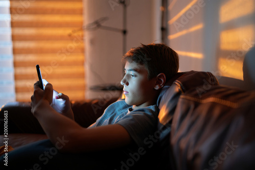 Teenage boy sitting on couch using smartphone in modern living room, warm evening light and authentic lifestyle photography