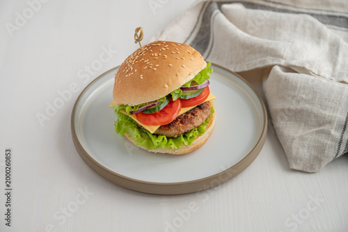 Beef burger with tomato, cheese, and lettuce on a plate on a light wooden background with napkin.
