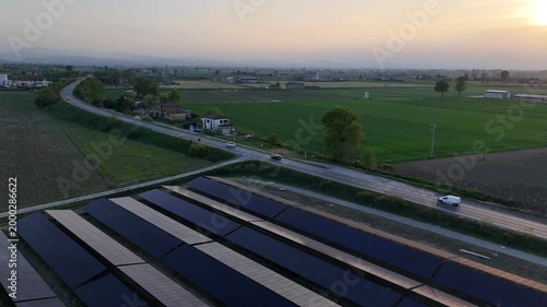 Aerial view of a large solar farm under construction in the Po Valley at sunset, with the SS9 Via Emilia road visible nearby and agricultural fields stretching to the horizon