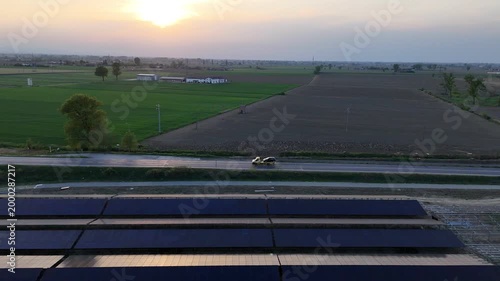 Aerial view of a large solar panel installation under construction in the Po Valley at sunset, with a road and passing vehicles visible