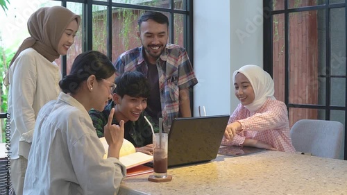 Asian friends gathered around laptop smiling as teenage student presents project to hijabwearing peers and mentor in bright cafe