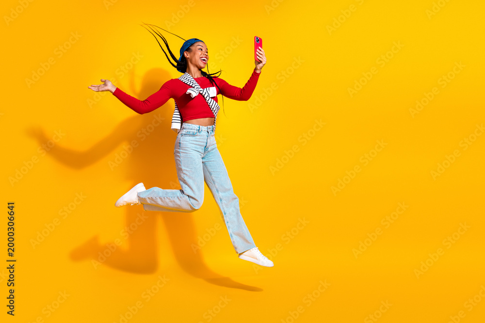 Fototapeta premium Joyful young woman jumping for a selfie against a bright yellow background