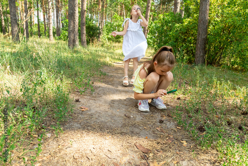 Two kids in casual clothes in summer exploring nature in forest together. Little girl looking through magnifying glass while squatting on ground