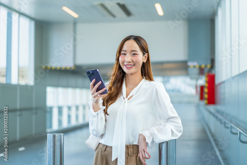 Fotografie Asian Businesswoman holding smartphone and smiling at camera in airport walkway