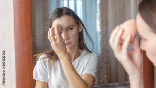 Tablou pe pânză Young woman with problems skin applying cosmetic foundation with applicator near mirror indoors