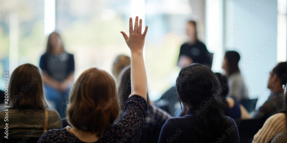 Obraz premium Participant raising hand to ask a question in sunlit seminar with presenters softly out of focus, modern corporate setting