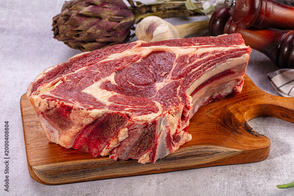 Obraz premium Raw beef rib eye steak on wooden board. Fresh artichoke, garlic, bay leaves, green onions, pepper mills. Gourmet cooking ingredients
