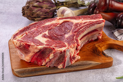 Raw beef rib eye steak on wooden board. Fresh artichoke, garlic, bay leaves, green onions, pepper mills. Gourmet cooking ingredients