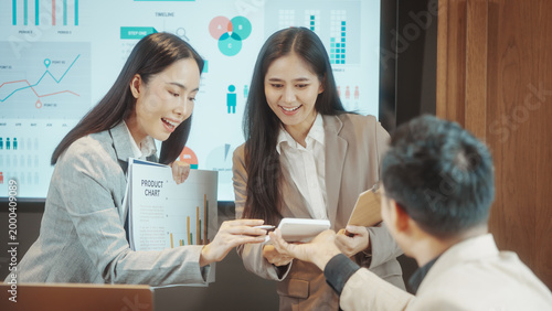 Smiling businesswoman presenting a printed bar chart in a meeting room with analytics dashboard on screen, colleagues listening and raising hand; modern corporate training, data-driven presentation.