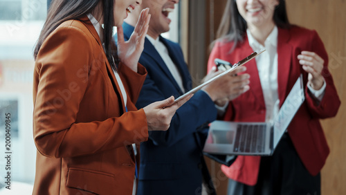 Business colleagues laughing and chatting in office corridor; woman in orange blazer holds clipboard, others with laptop and phone, teamwork and friendly corporate meeting.