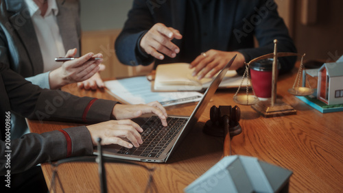 Business meeting at wooden table showing hands, laptop, legal gavel and scales of justice, documents and model house, warm lighting