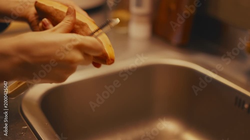 Close-up shot of female hands peeling raw sweet potato with a peeler over a kitchen sink. Warm lighting, selective focus with blurred metal countertop and wooden cutting board in the background.