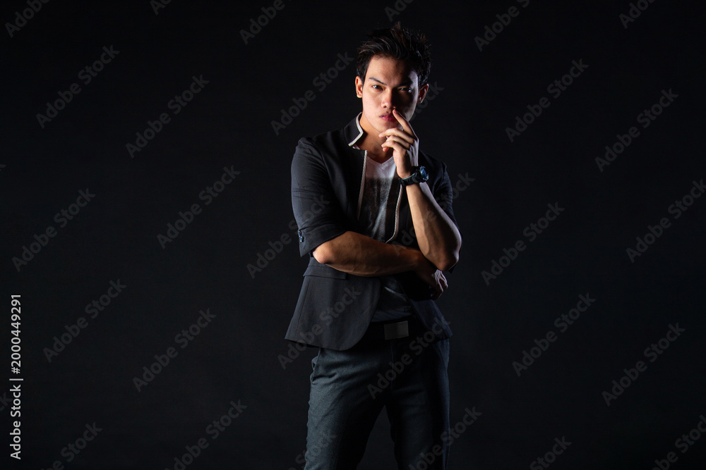 Fototapeta premium Young man poses confidently against dark studio backdrop, with dramatic lighting setup. Suit jacket layered over casual shirt, stylish yet edgy.