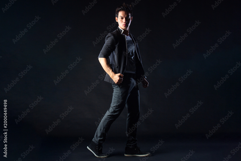 Fototapeta premium Young man poses confidently against dark studio backdrop, with dramatic lighting setup. Suit jacket layered over casual shirt, stylish yet edgy.