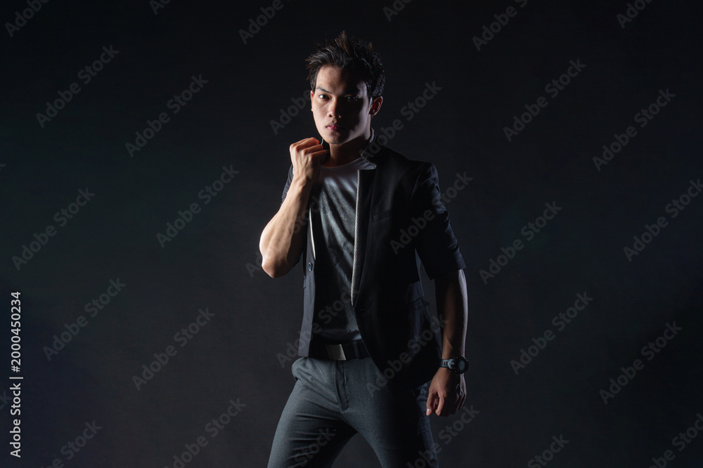 Obraz premium Young man poses confidently against dark studio backdrop, with dramatic lighting setup. Suit jacket layered over casual shirt, stylish yet edgy.