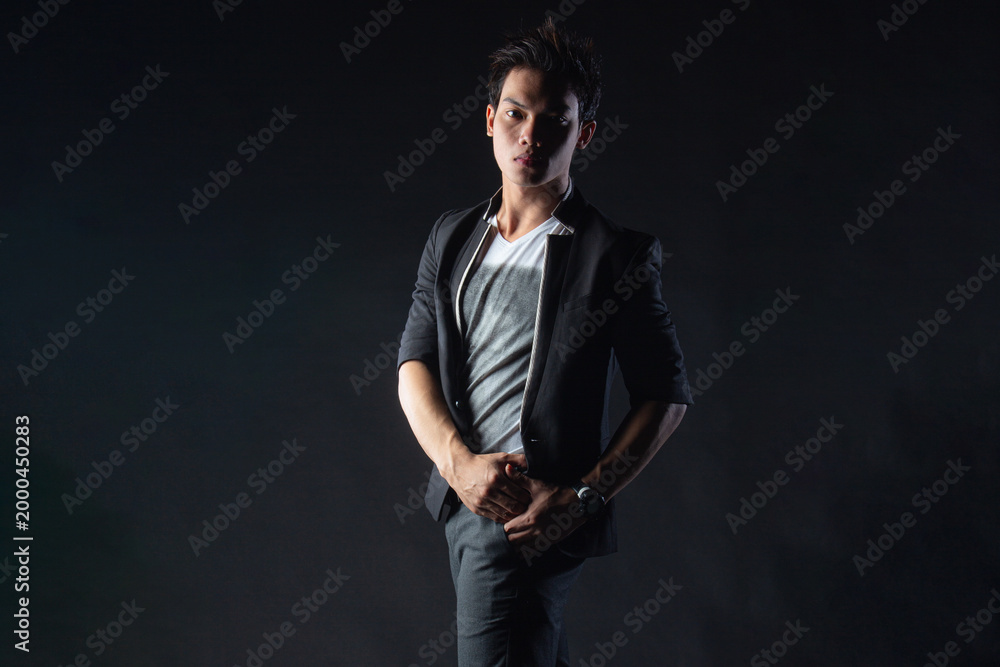 Fototapeta premium Young man poses confidently against dark studio backdrop, with dramatic lighting setup. Suit jacket layered over casual shirt, stylish yet edgy.