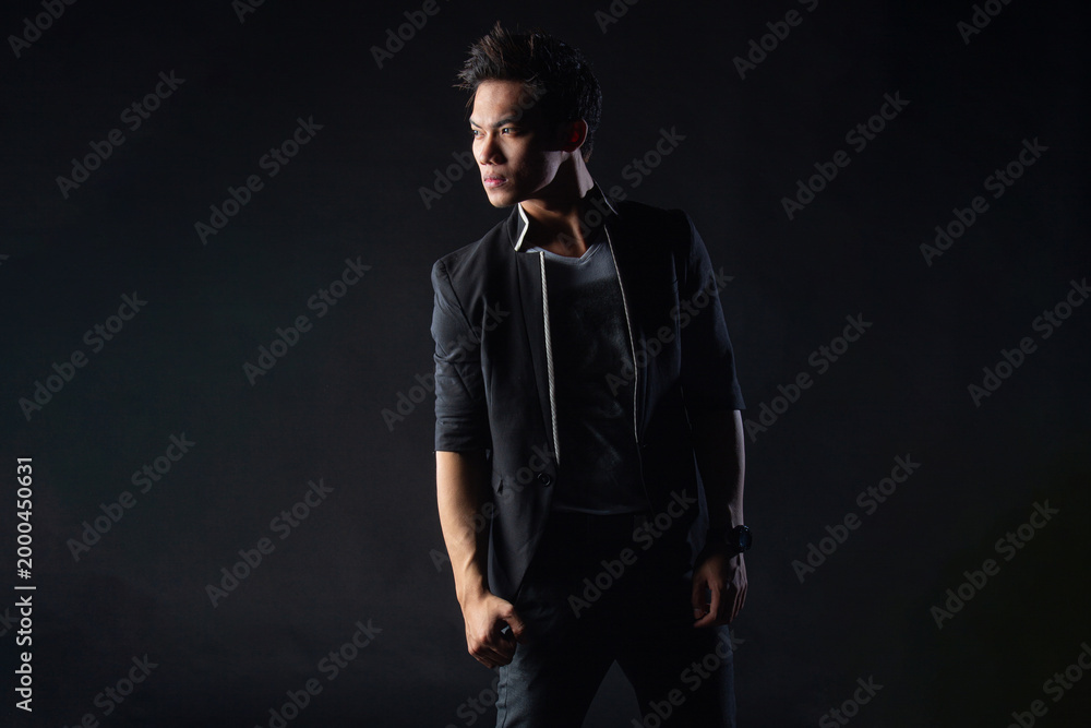 Fototapeta premium Young man poses confidently against dark studio backdrop, with dramatic lighting setup. Suit jacket layered over casual shirt, stylish yet edgy.