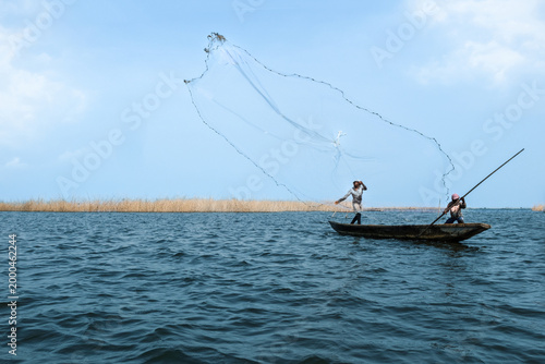 Ganvie, Benin - 14 April 2023: View of fishermen casting a net from a narrow boat on the shimmering waters, reeds providing a textured backdrop against the pale sky.